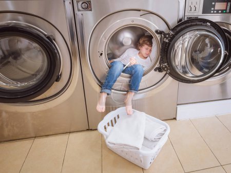 Téléchargez les photos : Corps complet de garçon pieds nus dormant dans le tambour de la machine à laver moderne dans la buanderie légère avec panier en plastique blanc - en image libre de droit