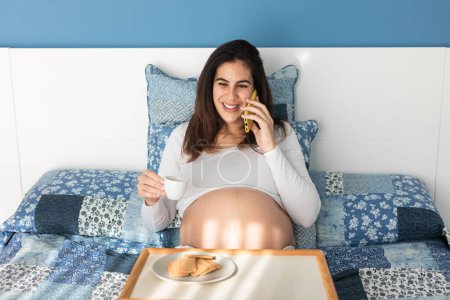 Photo for Glad pregnant female sitting on bed and talking on mobile phone while having coffee and cookies for breakfast - Royalty Free Image