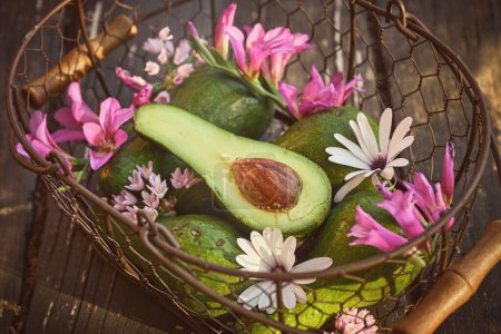 High angle of bunch of green avocado with pink and white flowers in metal basket placed on wooden table