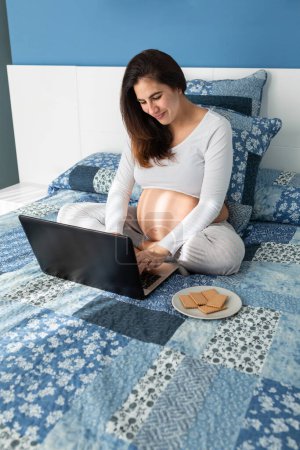 Photo for From above of smiling pregnant female browsing netbook while sitting on comfortable bed at home - Royalty Free Image