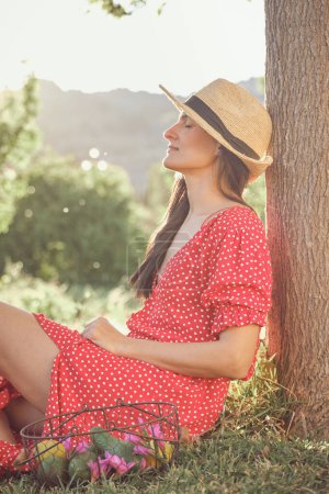 Téléchargez les photos : Vue latérale de la jeune femme en robe d'été et chapeau de paille assis sur l'herbe - en image libre de droit