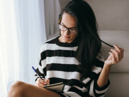 Téléchargez les photos : Attrayant femelle dans des lunettes élégantes prendre des notes dans un carnet tout en étant assis près du canapé blanc dans le salon de la lumière à la maison - en image libre de droit