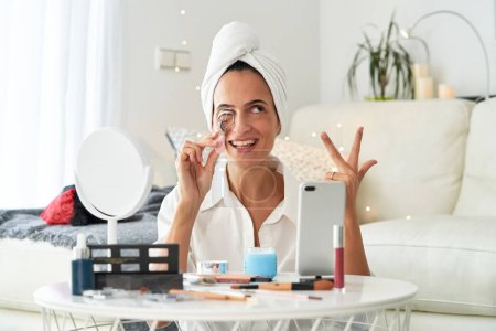 Téléchargez les photos : Blogueuse joyeuse avec serviette sur la tête en utilisant un bigoudi des cils tout en faisant du maquillage et en enregistrant un tutoriel de beauté sur téléphone portable à la maison et en regardant ailleurs en comptant le temps d'arrêt - en image libre de droit
