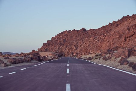Foto de Autopista recta de asfalto con líneas de marcado que van entre la cordillera rocosa áspera contra el cielo azul en la naturaleza en el día de verano - Imagen libre de derechos