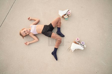 Foto de Desde arriba de niño sonriente en ropa deportiva acostado en el suelo del parque de skate cerca de quads con ruedas de colores mientras descansa después del entrenamiento - Imagen libre de derechos