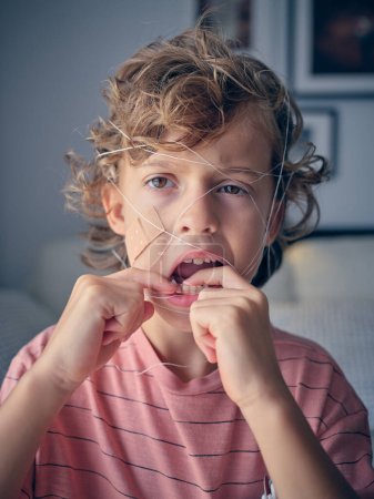 Photo for Brave boy in casual wear with thread on face looking away while touching loose tooth in light living room at home - Royalty Free Image
