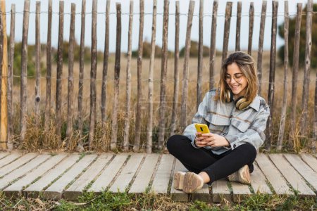 Téléchargez les photos : Jeune femme millénaire en vêtements décontractés élégants et lunettes assis sur la frontière de pierre navigation sur smartphone près de clôture en bois rustique et regardant la caméra - en image libre de droit