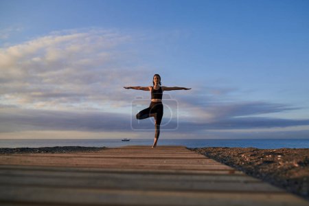 Téléchargez les photos : Corps entier de femme paisible faisant la pose de l'arbre avec les bras tendus tout en se tenant sur le sentier en bois près de la mer pendant l'entraînement de yoga dans la nature - en image libre de droit