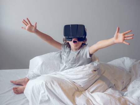 Photo for Amazed little barefoot kid wearing virtual reality goggles sitting on bed with raised arms and open mouth wrapped in blanket and experiencing cyberspace against gray background - Royalty Free Image