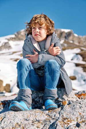 Photo for From below of blond kid with curly hair wearing warm outerwear sitting on rock near snowy mountain under blue sky and embracing himself while looking away - Royalty Free Image