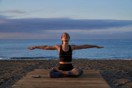 Téléchargez les photos : Corps entier de femme hispanique tranquille avec les yeux fermés pratiquant la posture Padmasana avec les bras écartés pendant la séance de yoga près de la mer - en image libre de droit