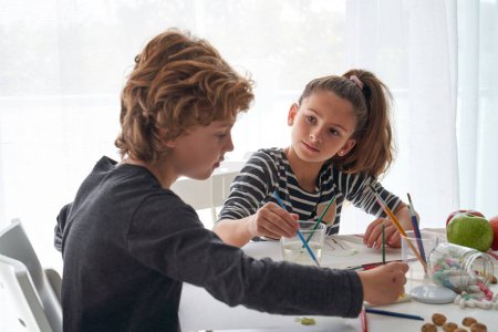 Téléchargez les photos : Adorables enfants peignant sur papier avec des pinceaux assis ensemble à table avec des pommes et des crayons éparpillés dans la pièce lumineuse - en image libre de droit
