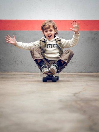 Photo for Full body of happy boy in hoodie sitting on skateboard with raised arms and opened mouth while having fun and looking at camera - Royalty Free Image