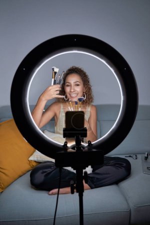 Photo for Full body of young female beauty vlogger demonstrating set of professional makeup brushes while recording video for vlog via smartphone and using tripod and ring lamp - Royalty Free Image