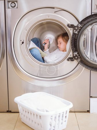 Photo for Side view of interested boy reading book with flashlight while lying in drum of modern washing machine near laundry basket - Royalty Free Image