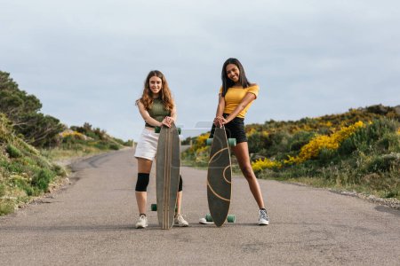 Photo for Full body of happy multiethnic girlfriends looking at camera other while walking on road with longboards - Royalty Free Image
