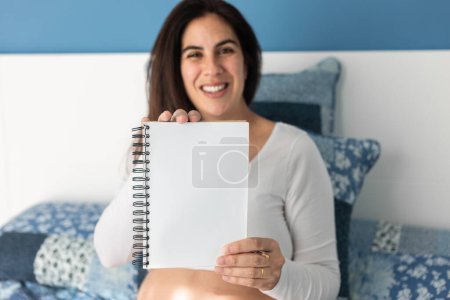 Photo for Delighted pregnant female sitting on soft bed at home and showing blank notepad at camera - Royalty Free Image