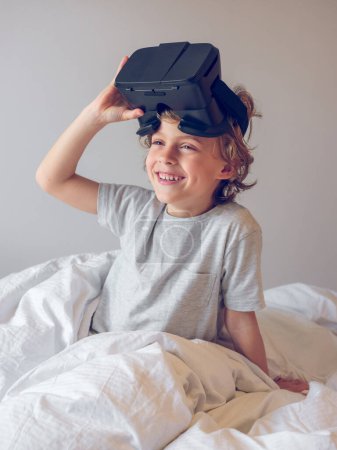 Photo for Cheerful little kid wearing virtual reality headset while sitting on bed under blanket and looking away against gray background at home - Royalty Free Image