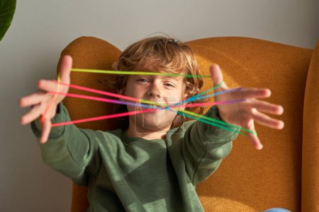 Photo for Satisfied preteen kid sitting in armchair and looking at camera while manipulating with colorful strings - Royalty Free Image
