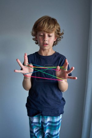 Photo for Portrait of surprised child in sleepwear playing with colorful finger thread and making string figures on gray background - Royalty Free Image
