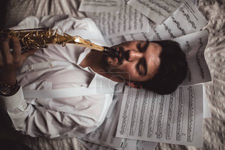 Téléchargez les photos : Jeune musicien allongé jouant du saxophone avec des partitions autour - en image libre de droit