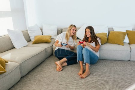 Téléchargez les photos : Corps complet d'amies positives avec des contrôleurs de jeu jouant au jeu vidéo assis sur le sol appuyé sur le canapé dans le salon à la maison - en image libre de droit