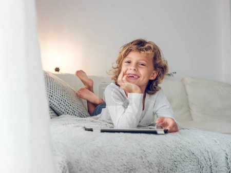 Photo for Full body of barefoot cheerful cute preteen boy lying on sofa with tablet and resting and looing away dreamily - Royalty Free Image