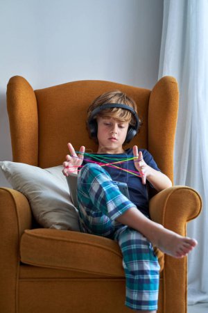 Foto de Niño enfocado en auriculares sentado en sillón acogedor con tableta en las rodillas y jugando con cuerda de dedo arco iris - Imagen libre de derechos