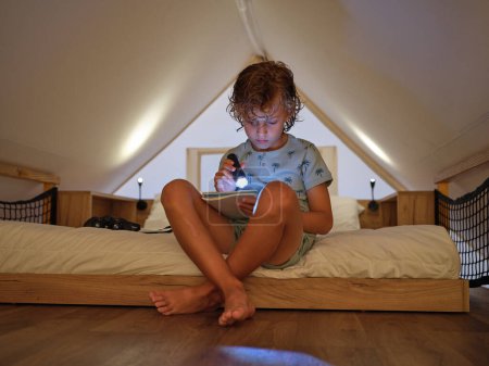 Photo for Full body of cute barefoot boy with glowing flashlight reading information on notebook while sitting on bed in light mansard - Royalty Free Image