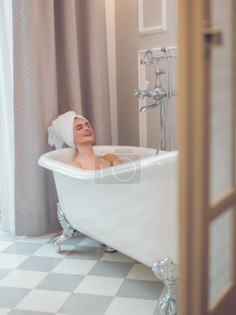 Photo for Young female with towel on head relaxing with closed eyes in soaker tub while smiling in modern bathroom - Royalty Free Image