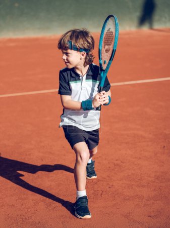 Foto de Cuerpo completo de adorable niño preadolescente enfocado en ropa deportiva preparándose para el derrame cerebral con raqueta mientras juega al tenis en pista dura - Imagen libre de derechos