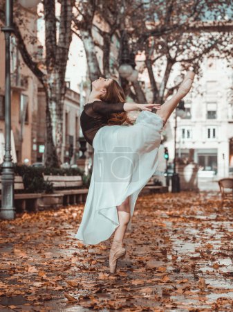 Foto de Vista lateral de la suave hembra en falda de ballet blanco de pie sobre hojas caídas con las piernas partidas en el patio de la ciudad - Imagen libre de derechos