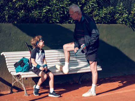 Téléchargez les photos : Vue latérale pleine longueur du tuteur chevelu gris senior avec raquette enseignant aux enfants souriants en vêtements de sport jouant au tennis assis sur un banc sur un terrain de sport par temps ensoleillé - en image libre de droit
