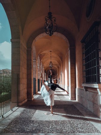 Foto de Cuerpo completo de esbelta bailarina elegante en traje de cuerpo y tutú de pie en arco salón del edificio con la pierna levantada en zapatos puntiagudos - Imagen libre de derechos