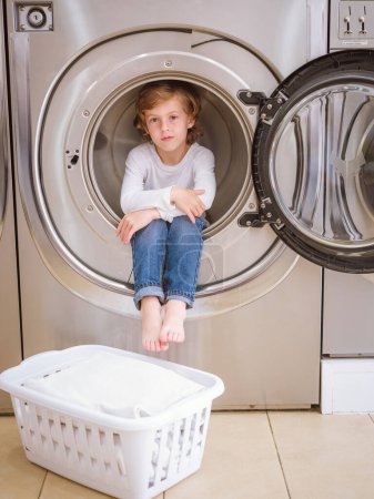 Foto de Cuerpo completo de adorable chico descalzo sentado en la bañera de la lavadora moderna y mirando a la cámara en el lavadero con cesta - Imagen libre de derechos