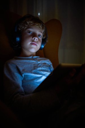 Foto de Stock de imagen vertical de un niño en auriculares sentados mirando una tableta por la noche con una mirada distraída. Tecnología - Imagen libre de derechos
