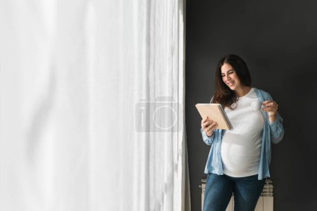 Photo for Cheerful pregnant female with cup of drink and notepad standing in room at home - Royalty Free Image