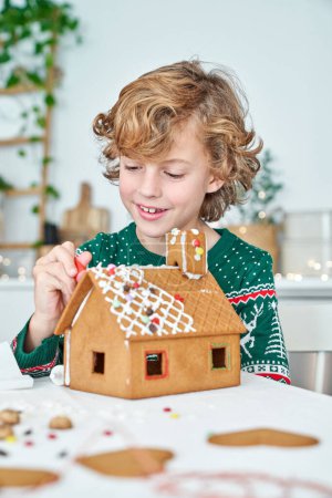 Photo for Positive boy gluing details of gingerbread house with sweet icing while sitting at table in light kitchen during Christmas preparation - Royalty Free Image