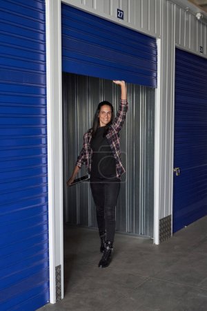 Photo for Full body of positive female in casual clothes with tablet in hand looking away while opening roll up door of metal storage - Royalty Free Image