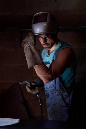 Foto de Vista lateral del soldador masculino barbudo en casco protector y guantes en taller preparándose para soldar con pistola de soldadura y mirando a la cámara - Imagen libre de derechos