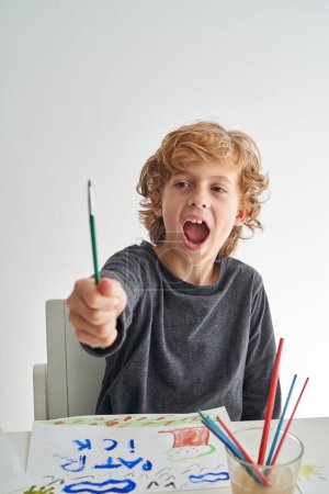 Photo for Amazed boy with opened mouth looking at paintbrush in outstretched arm while sitting at table with colorful drawing in room - Royalty Free Image