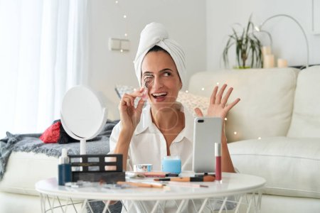 Foto de Mujer adulta feliz aplicando rizador de pestañas mientras graba video de maquillaje en línea para el blog de belleza en casa por la mañana y mirando hacia otro lado - Imagen libre de derechos