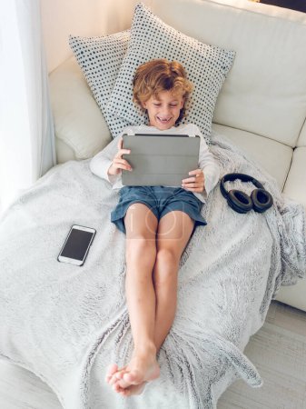 Photo for From above of full body barefoot cute preteen boy lying on soft plaid and cushion on sofa near smartphone and headphones and watching interesting video on tablet - Royalty Free Image