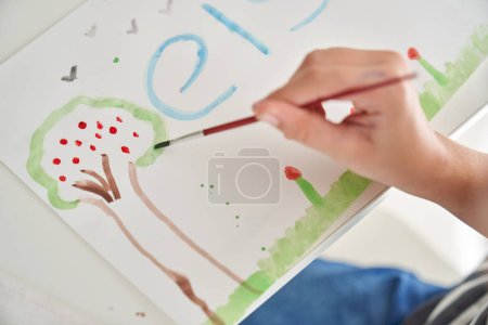 Photo for From above of crop anonymous kid with paintbrush drawing tree with colorful paint on white paper at table in light room - Royalty Free Image