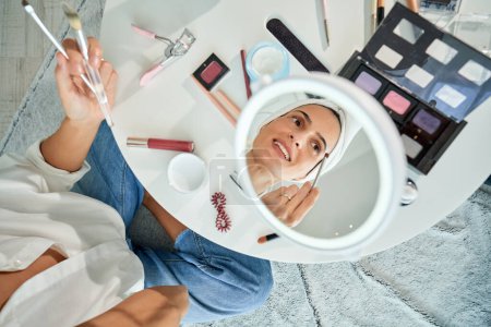 Photo for From above of reflection in mirror of positive female sitting at table and spreading eyeshadow with brush while using professional makeup palette - Royalty Free Image