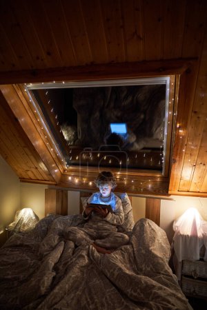 Téléchargez les photos : Garçon curieux en vêtements de nuit jouer au jeu sur tablette tout en étant couché sur le lit près de la fenêtre dans une chambre sombre avec guirlande lumineuse la nuit - en image libre de droit