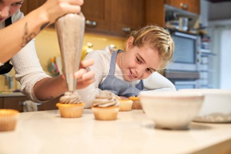 Téléchargez les photos : Preteen fille positive dans tablier admirant comment mère avec sac à pâtisserie décoré cupcakes au comptoir de la cuisine à la maison - en image libre de droit