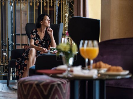 Téléchargez les photos : Jeune femme rêveuse assise à une table ronde avec dessert servi dans un restaurant de l'hôtel et regardant la fenêtre profiter de la vue sur la ville - en image libre de droit