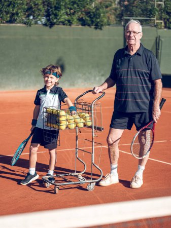 Photo for Full body of positive senior male coach and adorable preteen boy in sportswear and with tennis racket standing near balls cart on hard court in sunny day - Royalty Free Image
