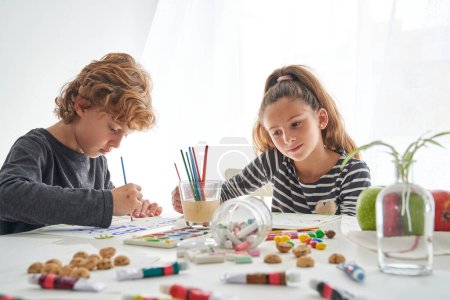 Téléchargez les photos : Enfants focalisés peignant avec des peintures colorées tout en étant assis ensemble à table avec un verre d'eau et diverses fournitures dispersées dans la salle de lumière - en image libre de droit